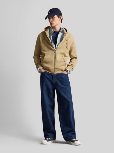 Męski model nosi Regular Fit Sweatjacke aus Baumwoll-Mix von Polo Ralph Lauren w beżowym kolorze, niebieskie szerokie dżinsy, biało-granatowe sneakersy, jasnoniebieską koszulę i granatową czapkę z daszkiem, całość w stonowanych kolorach, gładkie tekstury, Ganzkörperansicht von vorne.