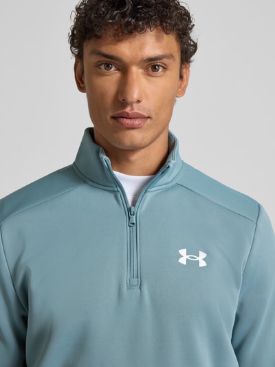 Männermodel trägt Sweatshirt mit Label-Detail von Under Armour in Blau, Stehkragen, kurzer Reißverschluss, Nahaufnahme Oberkörper. Lockerer Schnitt, gerippte Abschlüsse, Rückenlänge bei Größe M 72 cm, Ärmellänge 67 cm.