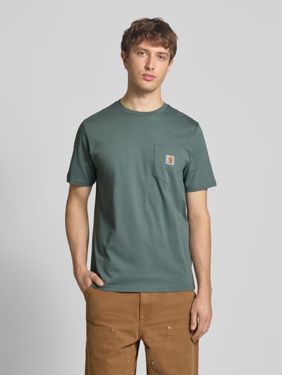 Männermodel trägt Regular Fit T-Shirt mit Logo-Patch von Carhartt Work In Progress in Grün, reine Baumwolle, Rundhals, Brusttasche, Ganzkörperansicht von vorne.