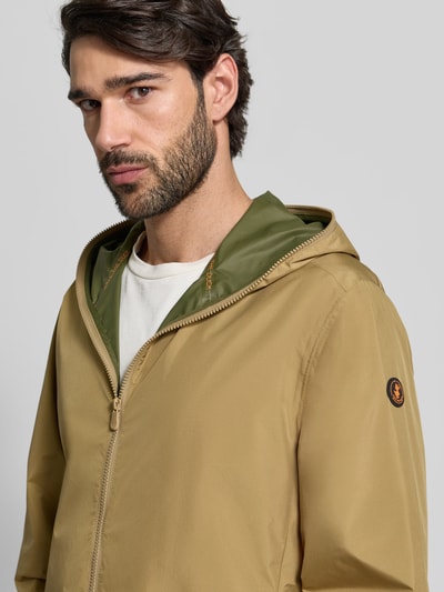 Männermodel trägt Jacke mit Zweiwegereißverschluss Modell 'Zayn' von SAVE THE DUCK in Beige mit olivefarbener Kapuze, Nahaufnahme Oberkörper, Regular Fit, sichtbares Markenlogo, glatte Oberfläche.