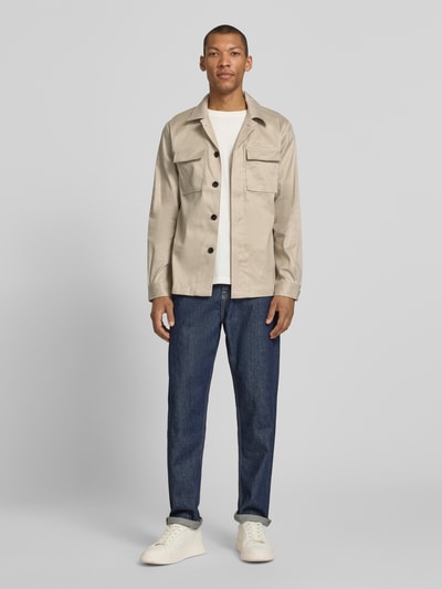 Männermodel trägt Regular Fit Hemdjacke aus Leinen-Mix mit Brusttaschen von Profuomo in Beige mit Reverskragen, verdeckter Knopfleiste, kombiniert mit blauen Jeans, glatte Struktur, Ganzkörperansicht von vorne.
