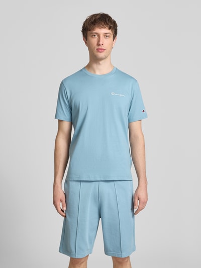 Mężczyzna nosi T-Shirt mit Label-Stitchings und Rundhalsausschnitt von CHAMPION w kolorze jasnoniebieskim, krój regular fit, czysta bawełna, przód z lekką fakturą, długość tyłu w rozmiarze M: 69 cm, widok całej sylwetki z przodu.