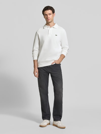 Männermodel trägt weißes Regular Fit Sweatshirt aus reiner Baumwolle von Lacoste zu dunkler Jeans, Ganzkörperansicht von vorne. Herren Sweatshirt von Lacoste