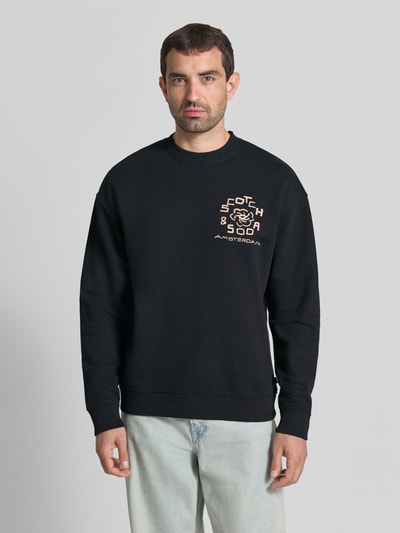 Männermodel draagt Relaxed Fit Pullover met logo-print van Scotch & Soda in zwart, puur katoen, ronde hals, elastische boordjes, Ganzkörperansicht von vorne.