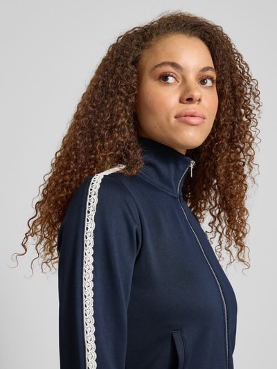 Vrouwenmodel draagt Regular Fit Trackjacke met Logostitching van Review in donkerblauw met opstaande kraag, met kant, ritssluiting, steekzakken, elastische boorden, detailopname bovenlichaam