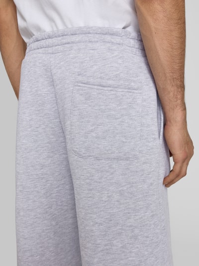Mężczyznamodel nosi Sweatshorts mit Logo-Print od REVIEW, mieszanka bawełny, krój loose fit, elastyczny pas, nadruk z logo, jasnoszary kolor, długość wewnętrzna nogawki 28 cm, szerokość w rozmiarze M: 45 cm, detaliczne zdjęcie z tyłu.