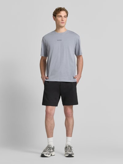 Męskimodel nosi Relaxed Fit T-Shirt aus reiner Baumwolle Modell 'TEE 10' od BOSS Green, jasnoszary t-shirt, czarne szorty, biało-czarne sneakersy, gładka faktura, pełny widok z przodu.