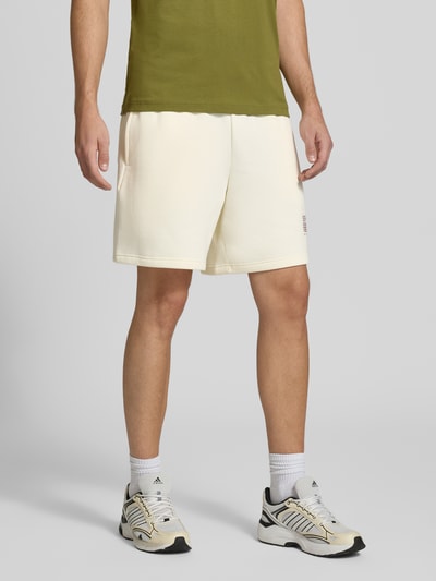 Männermodel trägt Sweatshorts aus Baumwoll-Mix von New Balance in Creme mit elastischem Bund, Label-Print und Gesäßtasche, Ganzkörperansicht von vorne.
