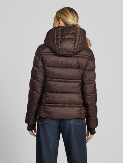 Frauenmodel trägt Steppjacke mit Kapuze und Reißverschlusstaschen von MICHAEL Michael Kors in Dunkelbraun, gesteppte Textur, Ganzkörperansicht Rückansicht. Modell 'PACKABLE WITH BIB', gerader Schnitt, Kapuze, seitliche Reißverschlusstaschen, elastische Abschlüsse mit Daumenloch.