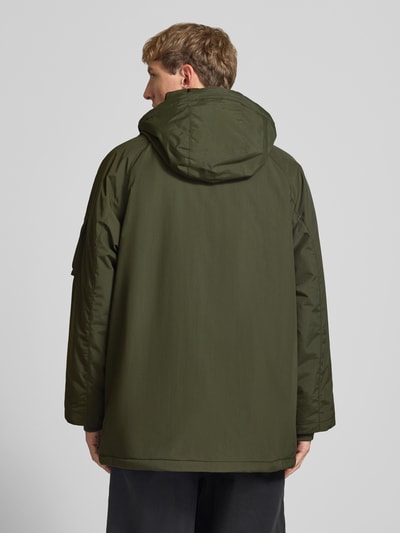 Männermodel trägt Parka mit Pattentaschen Modell 'Stanwood' von Carhartt Work In Progress in Olivgrün, Ganzkörperansicht Rückansicht. Regular Fit, Kapuze mit Tunnelzug, Ärmel-Reißverschlusstasche, Klettverschluss an Pattentaschen, Label-Patch, Reiß- und Klettverschluss, Maße für Größe M.