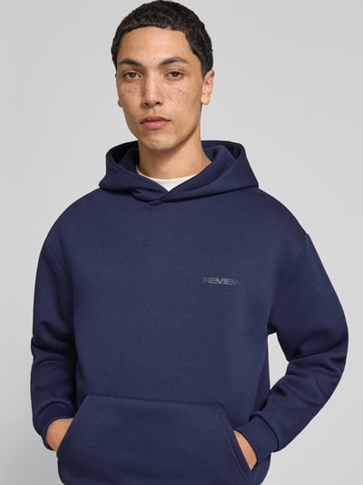 Männermodel trägt Hoodie mit Logo-Print von REVIEW in Dunkelblau, lockere Passform, Baumwoll-Mix, Kängurutasche, Nahaufnahme Oberkörper.