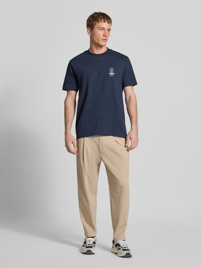 Męski model nosi spodnie z zakładkami w pasie Relaxed Fit Bundfaltenhose mit Viskose-Anteil Modell 'VIGGO CROPPED' von Marc O'Polo Denim, efekt: jednolity kolor, krój relaxed fit, średni stan, długość krótka, mieszanka materiałów z dodatkiem wiskozy, wpuszczane kieszenie, tylne kieszenie, tunel, elastyczny ściągacz, aplikacja z wyhaftowanym logo; widok całej sylwetki z przodu.