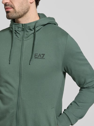 Männermodel trägt Slim Fit Sweatjacke mit Kapuze von EA7 Emporio Armani in Grün, Baumwoll-Mix, Logo-Print, Nahaufnahme Oberkörper.