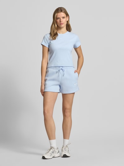 Frauenmodel trägt Sweatshorts mit Logo Modell 'Phoenix' von Nike aus Baumwoll-Mix, Regular-Fit, elastischer Bund mit Tunnelzug, Logo, hellblauer Farbton, glatte Textur, Bundweite bei Größe S 38 cm, Innenbeinlänge 10 cm, Ganzkörperansicht von vorne.