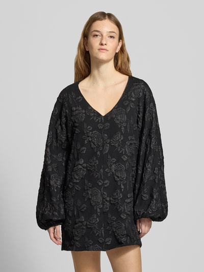 Vrouwenmodel draagt Minikleid mit V-Ausschnitt van Gina Tricot, zwart, losse fit, V-hals, sierbies, elastische mouwuiteinden, voorkant bovenlichaam close-up.