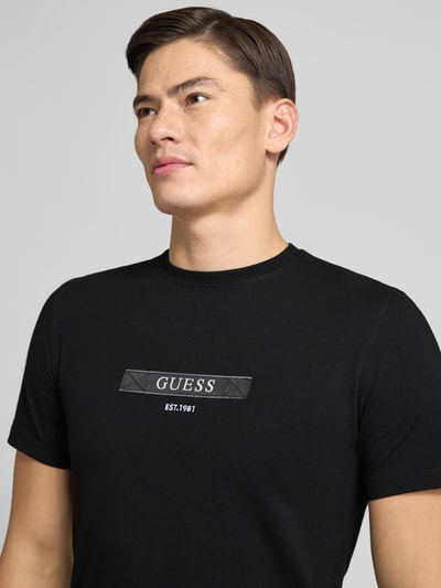 Männermodel trägt T-Shirt mit Label-Detail von Guess in Schwarz, Baumwoll-Elasthan-Mix, Regular-Fit, gerippter Rundhalsausschnitt, Nahaufnahme Oberkörper. Rückenlänge bei Größe M beträgt 68 cm.