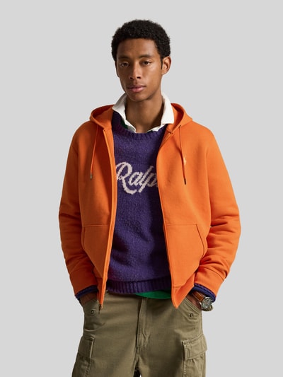 Mężczyznamodel nosi Regular Fit Sweatjacke aus Baumwoll-Mix von Polo Ralph Lauren w intensywnym pomarańczowym kolorze, prosty fason, gładka tekstura, widoczne kieszenie, tło szare, półzbliżenie na górną część ciała