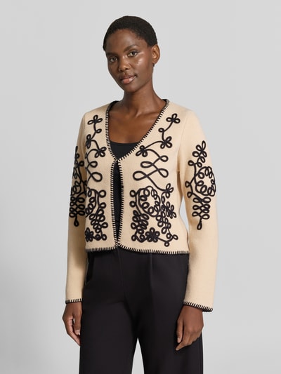 Modelka kobieca ma na sobie Strickjacke im Allover-Look mit Hakenverschluss od Apricot, beżowe i czarne kolory, ozdobne wzory, krój regular fit, dekolt w szpic, długość tyłu w rozmiarze S: 29 cm, długość rękawa: 63 cm, widok całej sylwetki z przodu.