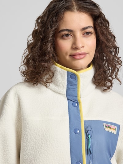 Frauenmodel trägt Fleecejacke mit Brusttasche und Label-Patch von Superdry in Weiß und Blau, gelber Akzent, Stehkragen, Druckknopfverschluss, Nahaufnahme Oberkörper, Kontext: Damen Fleecejacke, Gerader-Schnitt, Brusttasche mit Reißverschluss, Rückenlänge 57 cm, Schulterbreite 49 cm, Brustweite 61 cm, Ärmellänge 62 cm bei Größe S.