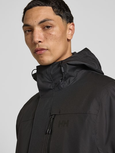 Männermodel trägt Regular Fit Jacke mit Logo-Print Modell 'Juell' von Helly Hansen in Schwarz, glatte Textur, Nahaufnahme Oberkörper.