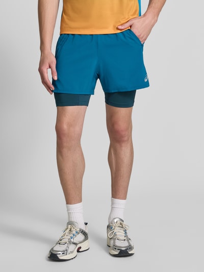 Mężczyzna nosi szorty Regular Fit Shorts z wbudowanymi legginsami Model 'Road' od ASICS TIGER w niebieskim kolorze, elastyczny pas, detal z logo, szerokość w rozmiarze M: 38 cm, długość wewnętrzna nogawki w rozmiarze M: 12 cm, krój regular fit, widok cała sylwetka z przodu.