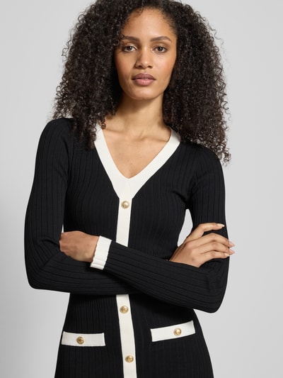 Vrouwenmodel draagt Slim Fit Tricotjurk uit viscosemix Model 'Laurel' van Guess, zwart met witte details, V-hals, goudkleurige knoopsluiting, opgestikte zakken, langmouw, context: Dames tricotjurk, ruglengte 110 cm, borstwijdte 33 cm, mouwlengte 63 cm. Nahaufnahme Oberkörper.