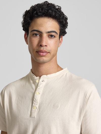 Männermodel trägt T-Shirt mit kurzer Knopfleiste von Superdry, cremefarben, Baumwoll-Mix mit Leinen-Anteil, Logo-Stitching, Nahaufnahme Oberkörper.
