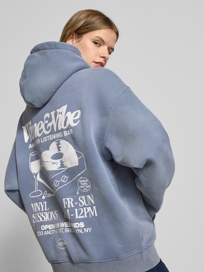 Kobiece model prezentuje Oversized Hoodie mit Känguru-Tasche Model 'Wine & Vibe' od ANOTHER COTTON LAB, mieszanka bawełny, efekt znoszenia, nadruk z logo, kieszeń kangurka, styl oversized, widok na całe plecy.