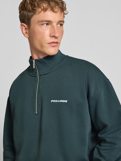Męski model nosi Relaxed Fit Sweatshirt aus Baumwoll-Mix od PEGADOR w ciemnozielonym kolorze, wyhaftowane logo, ozdobne szwy, prążkowane wykończenia, kołnierz zapinany, długi rękaw, mieszanka bawełny, długość tyłu w rozmiarze M: 72 cm, długość rękawów w rozmiarze M: 65 cm, zbliżenie na górną część ciała.