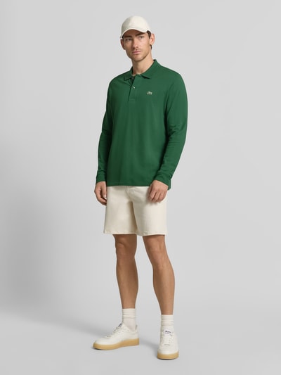 Männermodel trägt Regular Fit Poloshirt aus reiner Baumwolle von Lacoste in Grün, kombiniert mit heller Shorts, Ganzkörperansicht von vorne. Herren Poloshirt von Lacoste