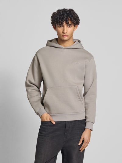 Männermodel trägt Hoodie mit Känguru-Tasche von REVIEW in Beige, lockere Passform, Label-Detail, elastische Abschlüsse, Ganzkörperansicht von vorne.