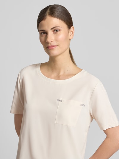 Kobiece model prezentuje damski t-shirt 'T-Shirt aus Viskose-Mix mit Brusttasche' od monari, mieszanka wiskozy z dodatkiem streczu, krój loose fit, okrągły dekolt, brustasza, obszycie z kamieni stras, detal z logo, długość tyłu w rozmiarze 36: 56 cm, jasny kolor, gładka tekstura, prosty krój, detaliczne zdobienie, zbliżenie na górną część ciała, półprofil, ujęcie z przodu