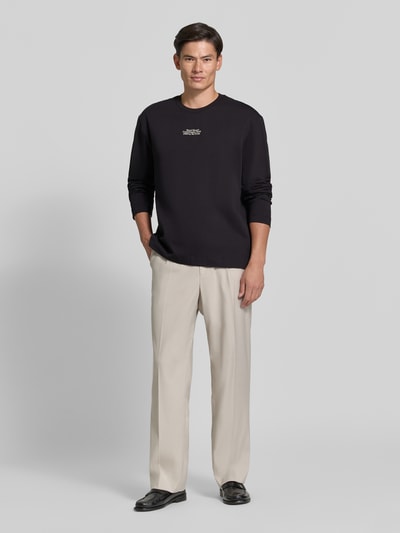 Männermodel trägt Regular Fit Longsleeve aus reiner Baumwolle Modell 'Ryan' von Wood Wood in Schwarz, Logo-Stitching, Ganzkörperansicht von vorne. Herren Longsleeve, Rückenlänge bei Größe M 79 cm, gerade beige Hose, glatte Textur.