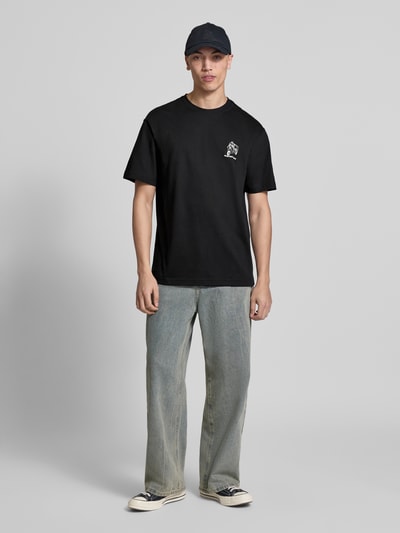 Männermodel trägt Relaxed Fit T-Shirt aus reiner Bio-Baumwolle Modell 'FRED' von Only & Sons, unifarben schwarz mit Schriftzug-Print, Rundhals und kurzen Ärmeln, kombiniert zu heller Jeans, Ganzkörperansicht von vorne.