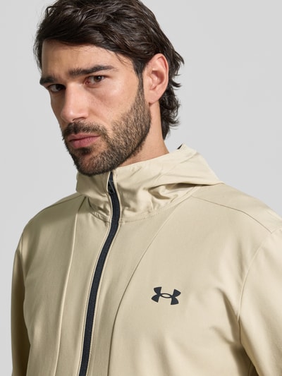 Männermodel trägt Regular Fit Jacke mit Logo-Print Modell 'Unstoppable' von Under Armour, Beige, Reißverschluss, Kapuze, Nahaufnahme Oberkörper.