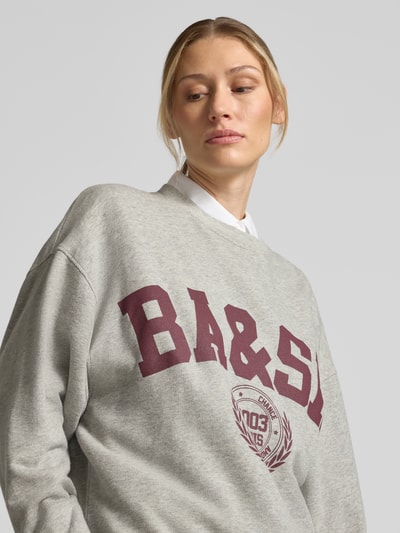 Kobiece modelka ma na sobie szarą bluzę ba&sh z nadrukiem z logo, mieszanka bawełny i elastanu, luźny krój, okrągły dekolt, Model 'BENJAMIN', nadruk z logo, obniżone ramiona, zbliżenie na górną część ciała.