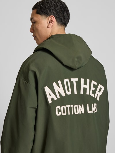 Męskki model nosi Adventure Jacke mit Kapuze und verstellbarem Saum von ANOTHER COTTON LAB, oliwkowa z nadrukiem z logo, prosty krój, naszywka z logo, długość tyłu w rozmiarze M: 70 cm, szerokość ramion w rozmiarze M: 60 cm, szerokość klatki piersiowej w rozmiarze M: 64 cm, długość rękawów w rozmiarze M: 53 cm, zapięcie na zatrzask, widok na całą sylwetkę z tyłu