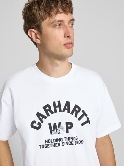 Męski model nosi Regular Fit T-Shirt aus reiner Baumwolle Model 'Column' von Carhartt Work In Progress w kolorze białym z nadrukiem z napisem i logo, okrągły dekolt, krój regular fit, długość tyłu w rozmiarze M: 75 cm, widok na górną część ciała z przodu.