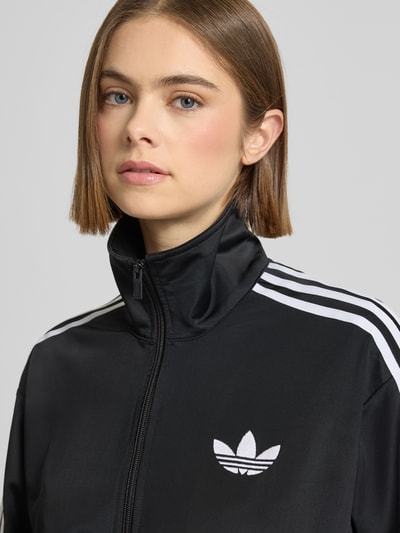 Frauenmodel trägt Sweatjacke mit Stehkragen von adidas Originals in Schwarz mit weißen Streifen, Logo-Label-Stitching, Reißverschluss, gerippte Abschlüsse, Reißverschlusstaschen, Nahaufnahme Oberkörper.