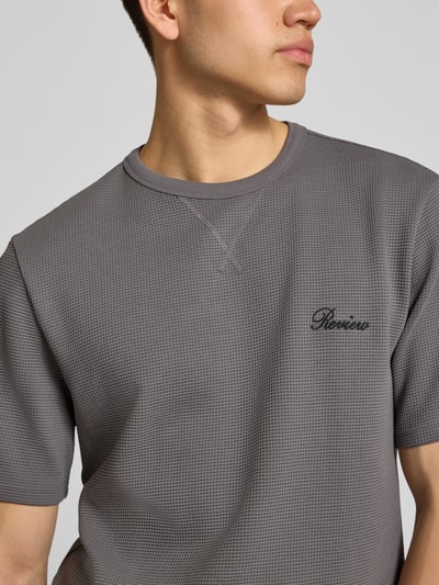 Männermodel trägt T-Shirt mit Label-Stitching von REVIEW in Grau mit Waffelstruktur und Rundhalsausschnitt, Nahaufnahme Oberkörper.