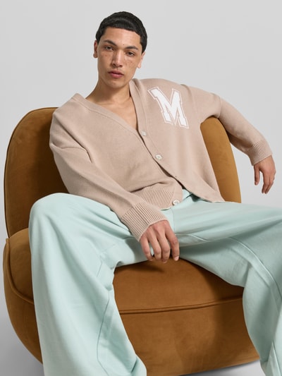 Männermodel trägt Oversized Cardigan aus Schurwoll-Mix von MM6 Maison Margiela in Strick-Optik, Beige mit M-Applikation, Ganzkörperansicht von vorne. Rückenlänge 78 cm, Schulterbreite 48 cm, Ärmellänge 69 cm, V-Ausschnitt, durchgängige Knopfleiste.
