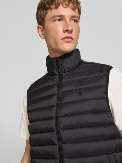 Männermodel trägt Steppweste mit Stehkragen und Label-Stitching von REVIEW, krój slim fit, zamek błyskawiczny, Długość tyłu w rozmiarze M: 68 cm, Szerokość ramion w rozmiarze M: 43 cm, Szerokość klatki piersiowej w rozmiarze M: 56 cm, czarny kolor, poziome pikowanie, gładka tekstura, Nahaufnahme górnej części ciała.