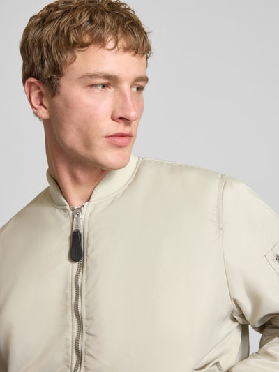 Männermodel trägt Regular Fit Bomberjacke mit Stehkragen Modell 'Heritage' von Alpha Industries, beige, glatte Textur, Nahaufnahme Oberkörper. Rückenlänge bei Größe M 63 cm, Schulterbreite 50 cm, Brustweite 57 cm, Reißverschluss, Pattentaschen, Elastische Abschlüsse.