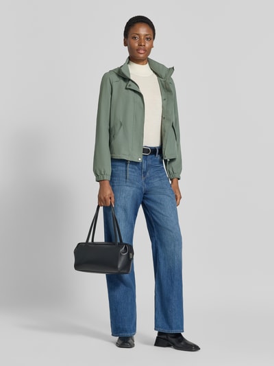 Frauenmodel trägt Regular Fit Parka mit Kapuzenkragen Modell 'ZOA' von VERO MODA in Salbeigrün, kombiniert mit Jeans und schwarzer Tasche, Ganzkörperansicht von vorne.