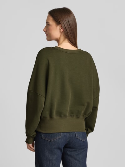 Kobiece modelka nosi Oversized Sweatshirt aus reiner Baumwolle von miss goodlife, kolor oliwkowy, krój oversized, okrągły dekolt, elastyczne wykończenia, nadruk z napisem, widok całej sylwetki z tyłu.