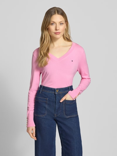 Vrouwenmodel draagt Regular Fit gebreide pullover uit puur katoen van Tommy Hilfiger in effen roze met gestikt logo, v-hals, lange mouw, sierknopen, geribde zoom, vooraanzicht van bovenlichaam.