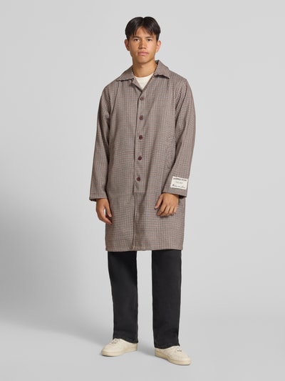 Männermodel trägt Trenchcoat mit Knopfleiste und Label-Patch von ANOTHER COTTON LAB, braun kariert, gerade geschnitten, Ganzkörperansicht von vorne.