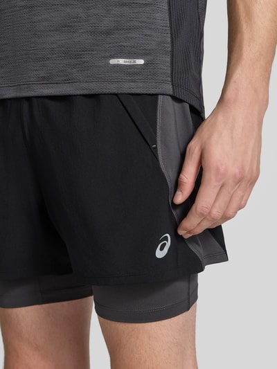 Mężczyznamodel nosi Regular Fit Shorts mit integrierten Tights Modell 'Road' von ASICS TIGER, czarne, detal z logo, krój regular fit, elastyczny pas, Szerokość w rozmiarze M: 38 cm, Długość wewnętrzna nogawki w rozmiarze M: 12 cm, Ganzkörperansicht z przodu, zdjęcie zbliżenie na górną część nóg.
