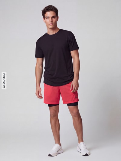 Männermodel trägt Relaxed Fit Sportshorts im Double-Layer-Look von Under Armour in Rot und Schwarz, Ganzkörperansicht von vorne, KI-modifiziertes Bild.