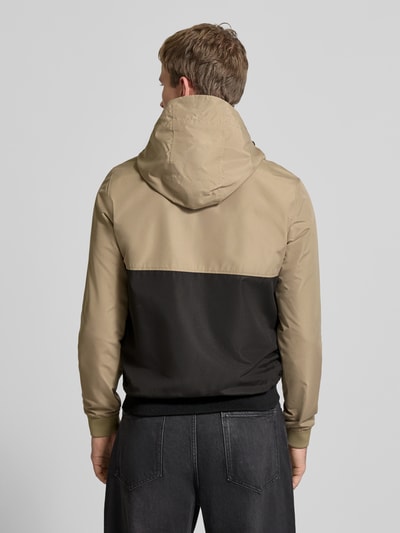 Männermodel trägt Jacke mit Kapuze und Reißverschluss Modell 'RUSH' von JACK & JONES in beige und schwarz, regular fit, mit tunnelkoord, logoprint, steekzakken, ruglengte bij maat M 68 cm, schouderbreedte 46 cm, borstwijdte 55 cm, mouwlengte 68 cm. Ganzkörperansicht Rückansicht.